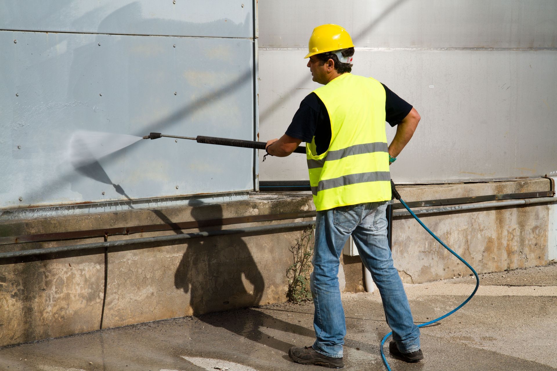 A construction worker in a high-visibility yellow vest and hard hat pressure-washing a metal industrial wall.