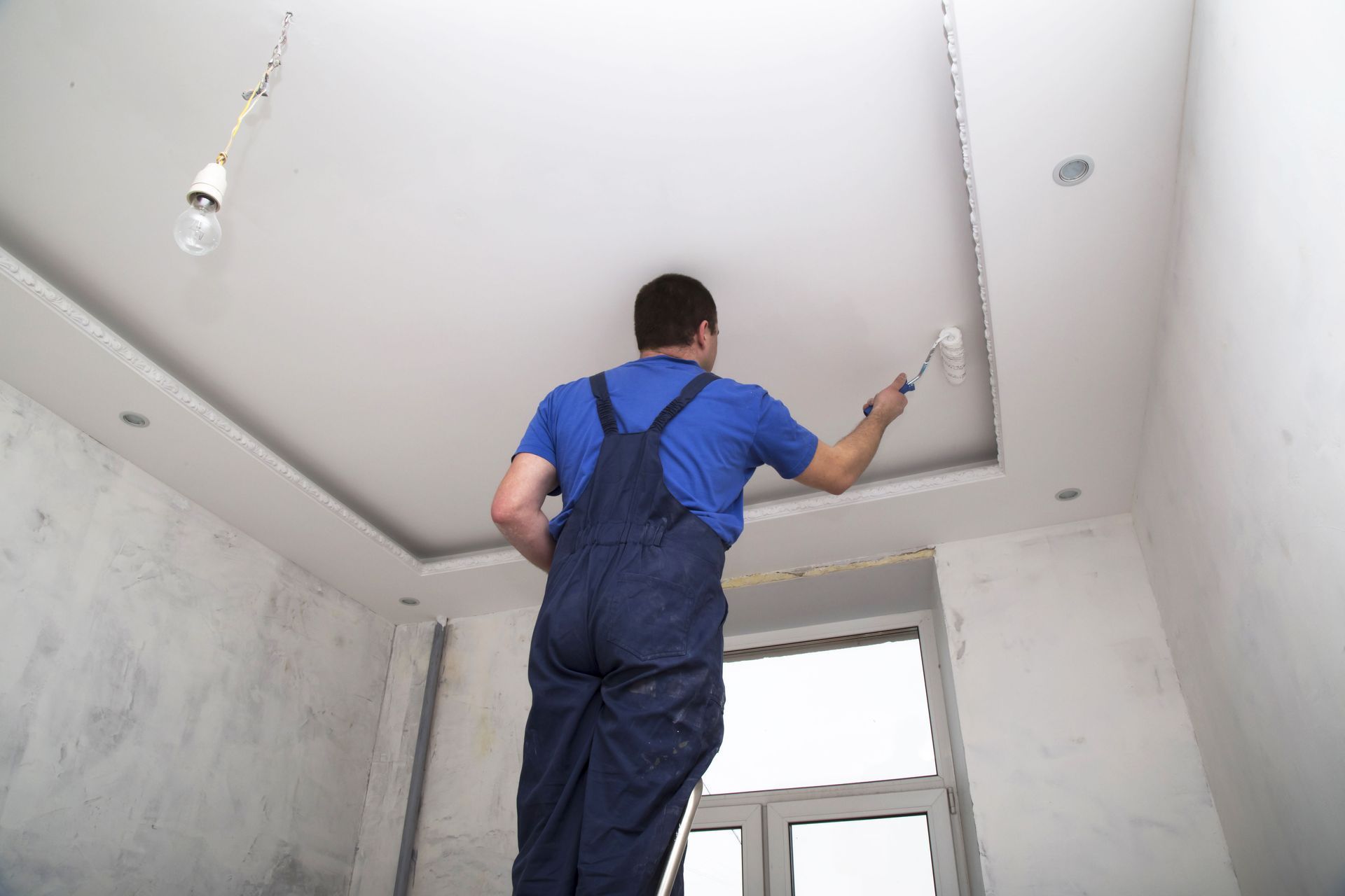 A person in blue coveralls stands on a ladder, painting a white ceiling in a room under construction.