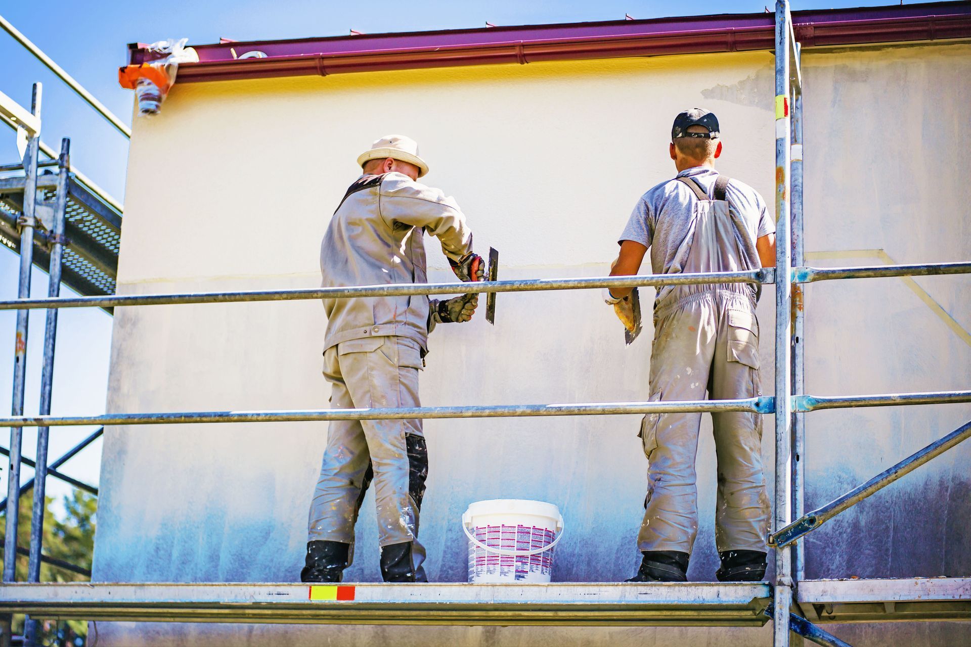 Two construction workers in work uniforms stand on scaffolding, smoothing plaster on an exterior building wall.