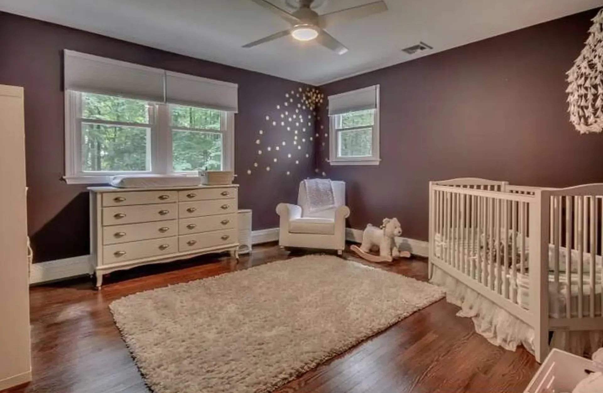 Nursery with crib, white dresser, rocking chair, and beige rug in a dark purple room