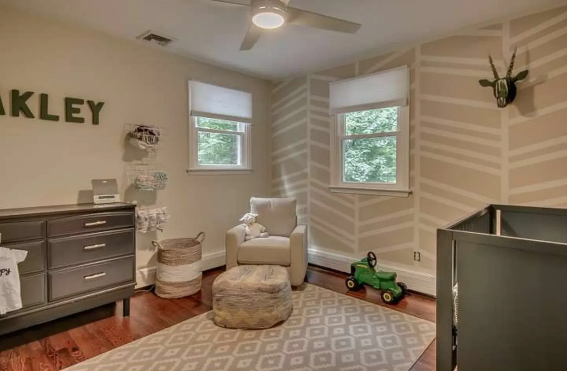 Neutral-toned nursery with crib, armchair, rug, dresser, and toy truck by the windows