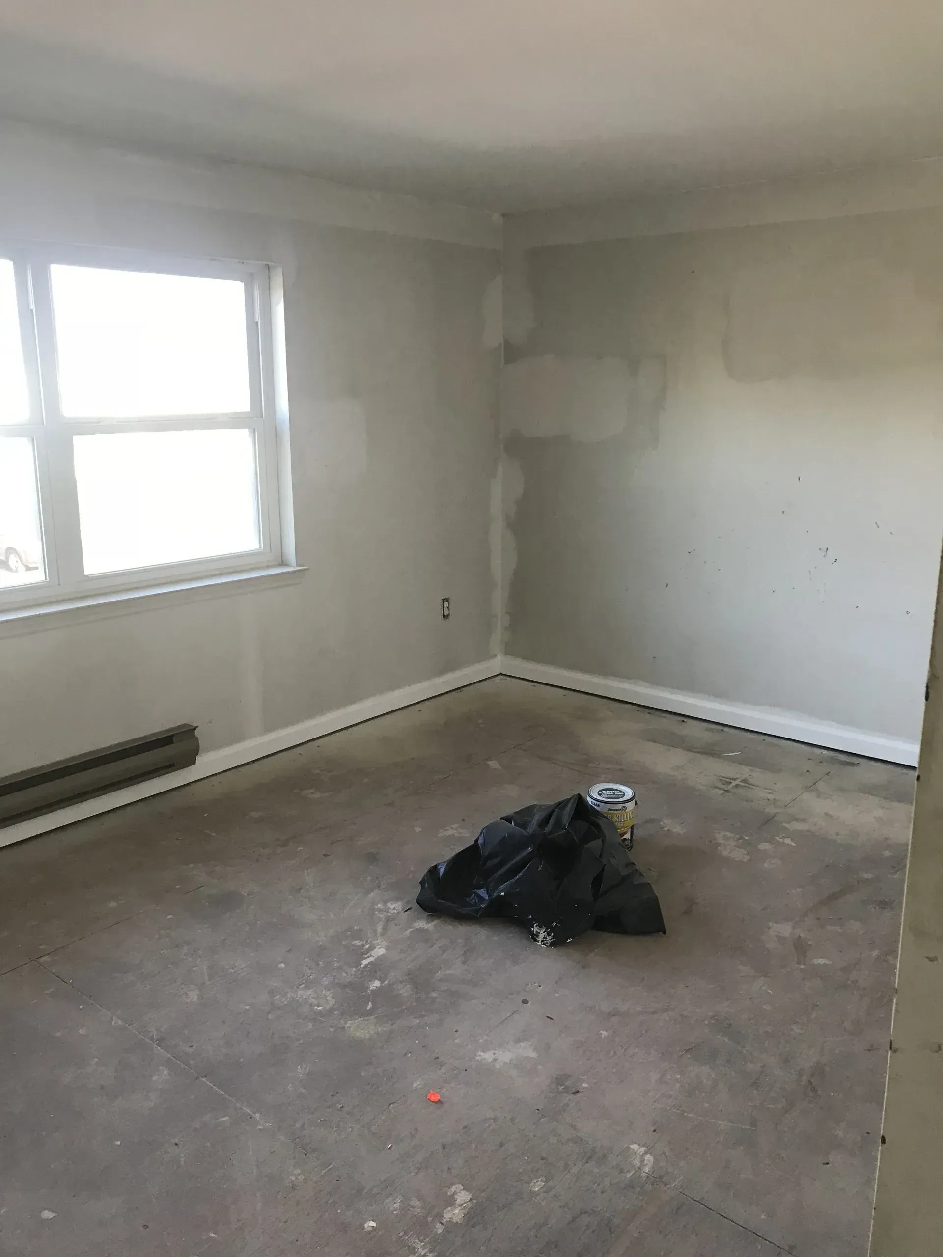 Empty room with white walls, a window, and a black trash bag on a bare concrete floor