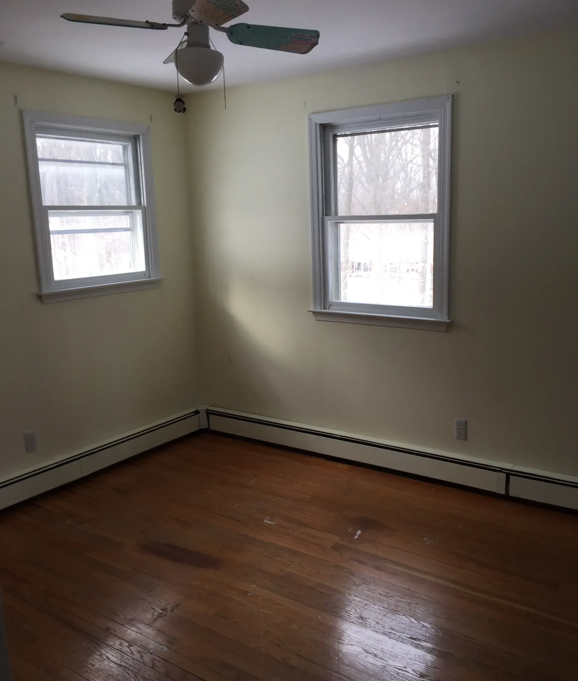 Empty room with hardwood floor, two windows, white walls, and a ceiling fan.