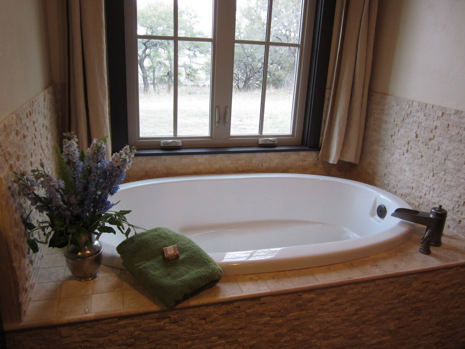 Oval bathtub next to a window, with a vase of flowers and a green towel on the side.