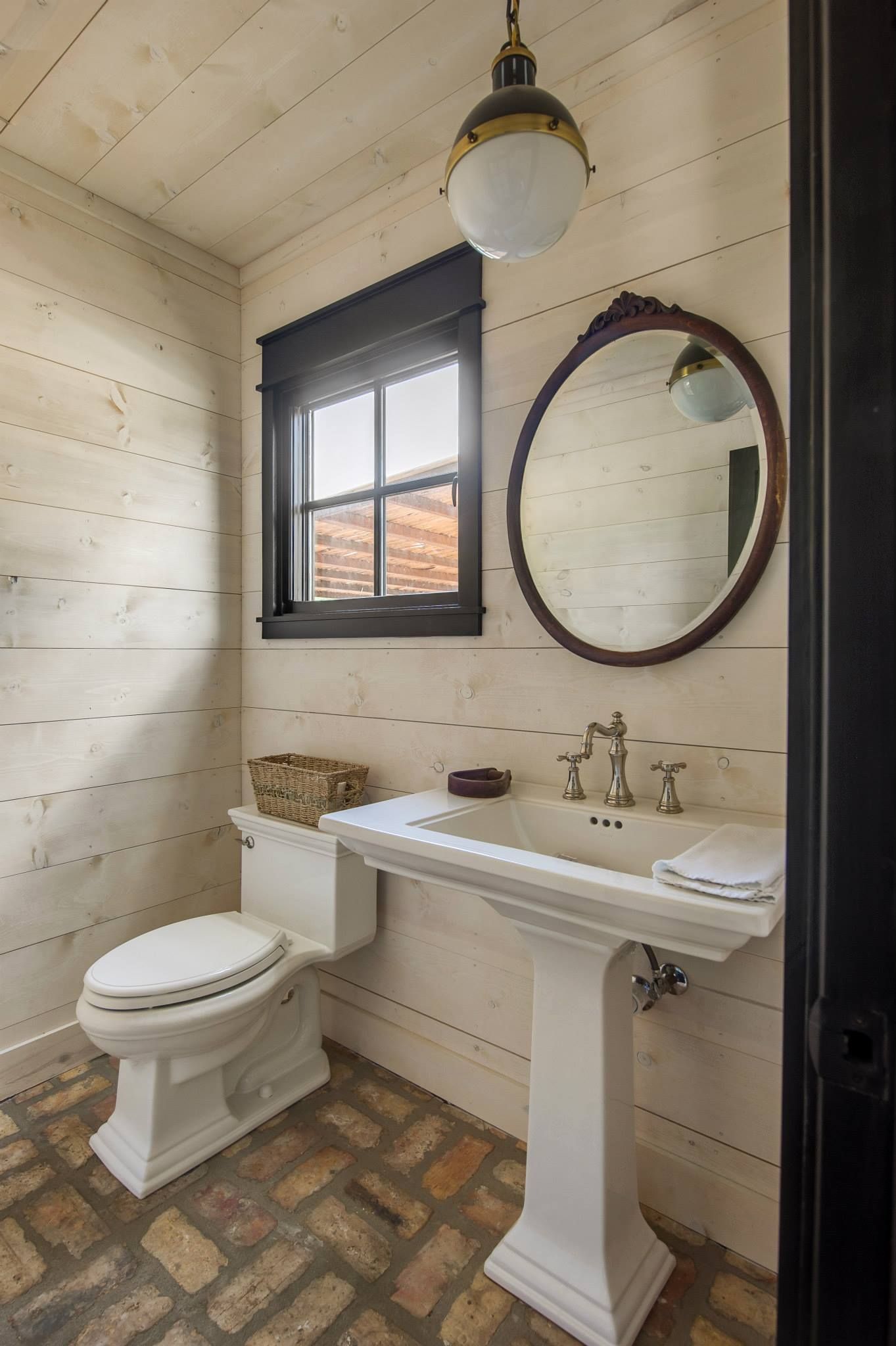 Small bathroom with a white pedestal sink and toilet, brick floor, wood walls, and a round mirror.