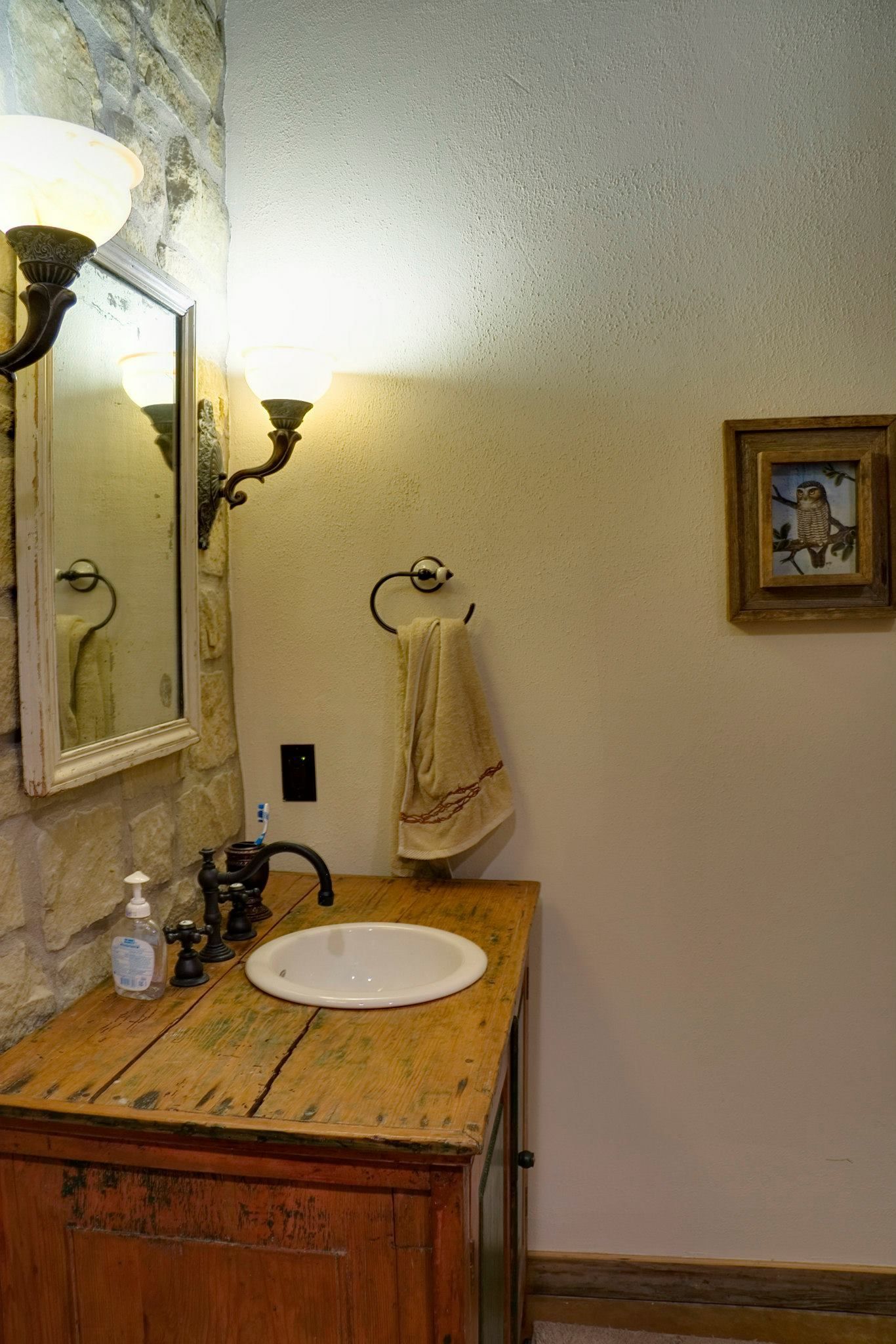 Rustic bathroom with wooden vanity, oval sink, mirror, wall sconces, and a framed photo.