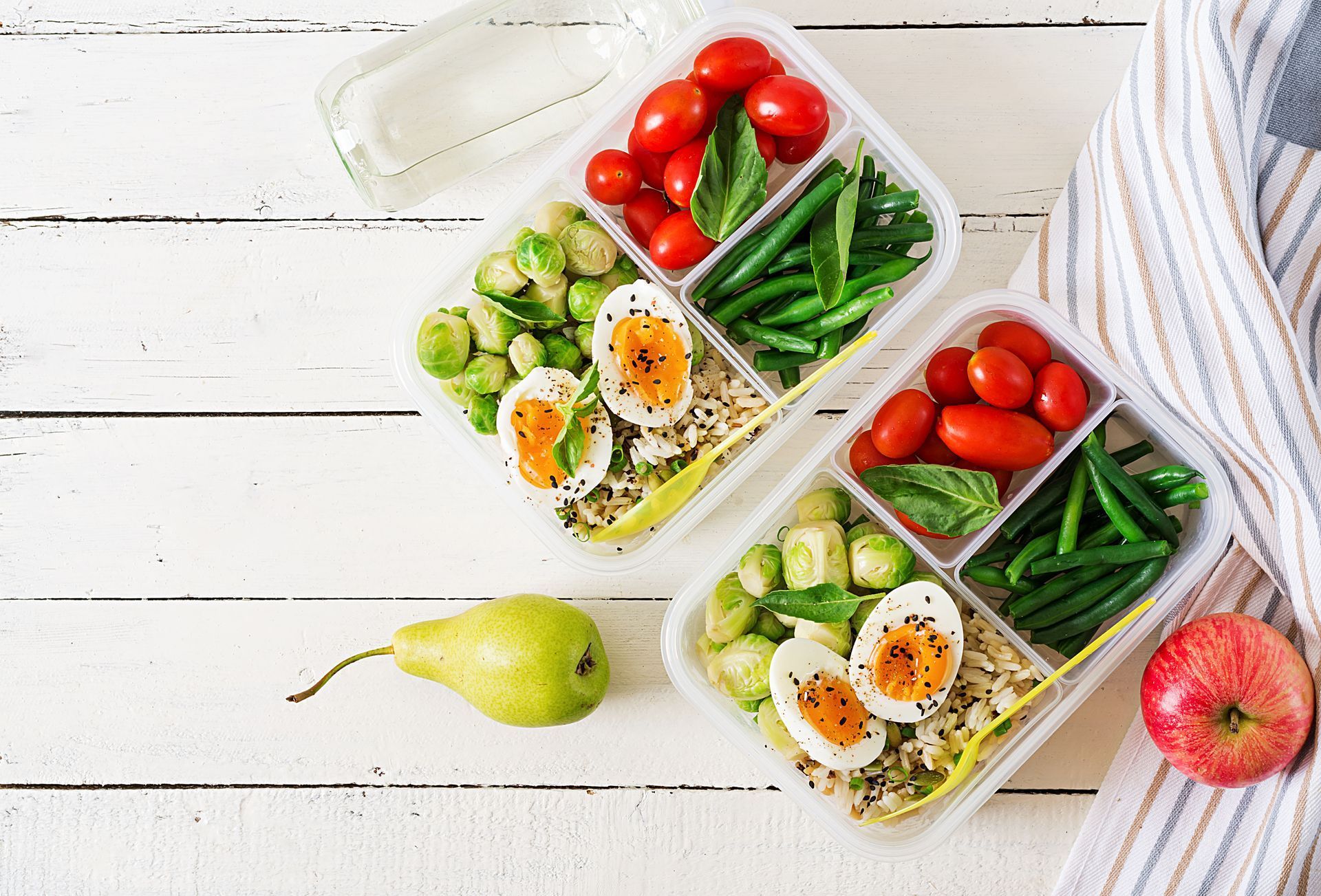 Lunch containers with tomatoes, green beans, Brussels sprouts, eggs, and grains, next to fruit.