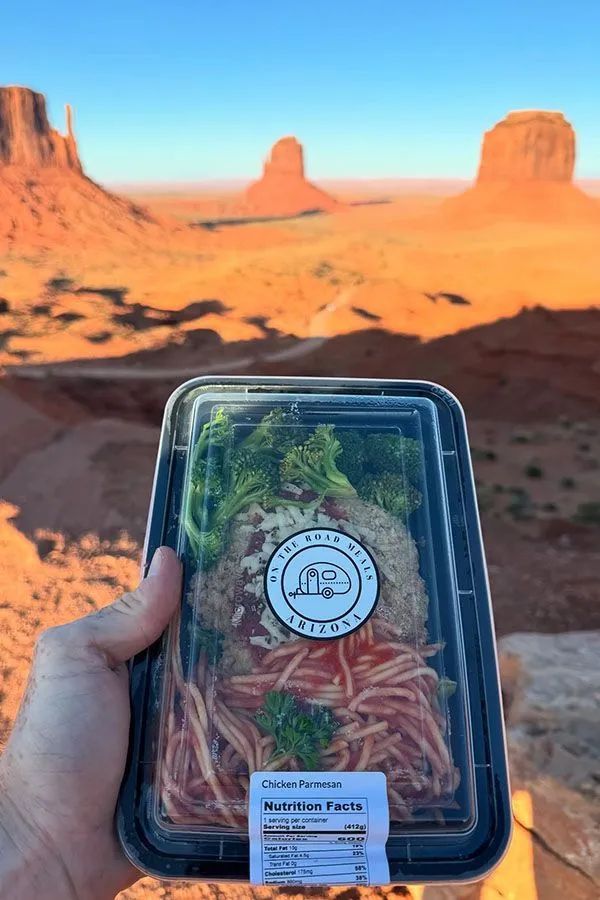 Hand holding a pre-packaged meal with red rock formations in the background. Sunny outdoor setting.