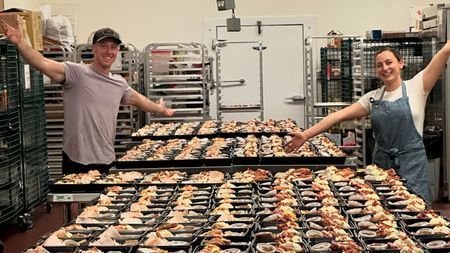 Two people with arms outstretched, standing in front of many trays of prepared food in a commercial kitchen.