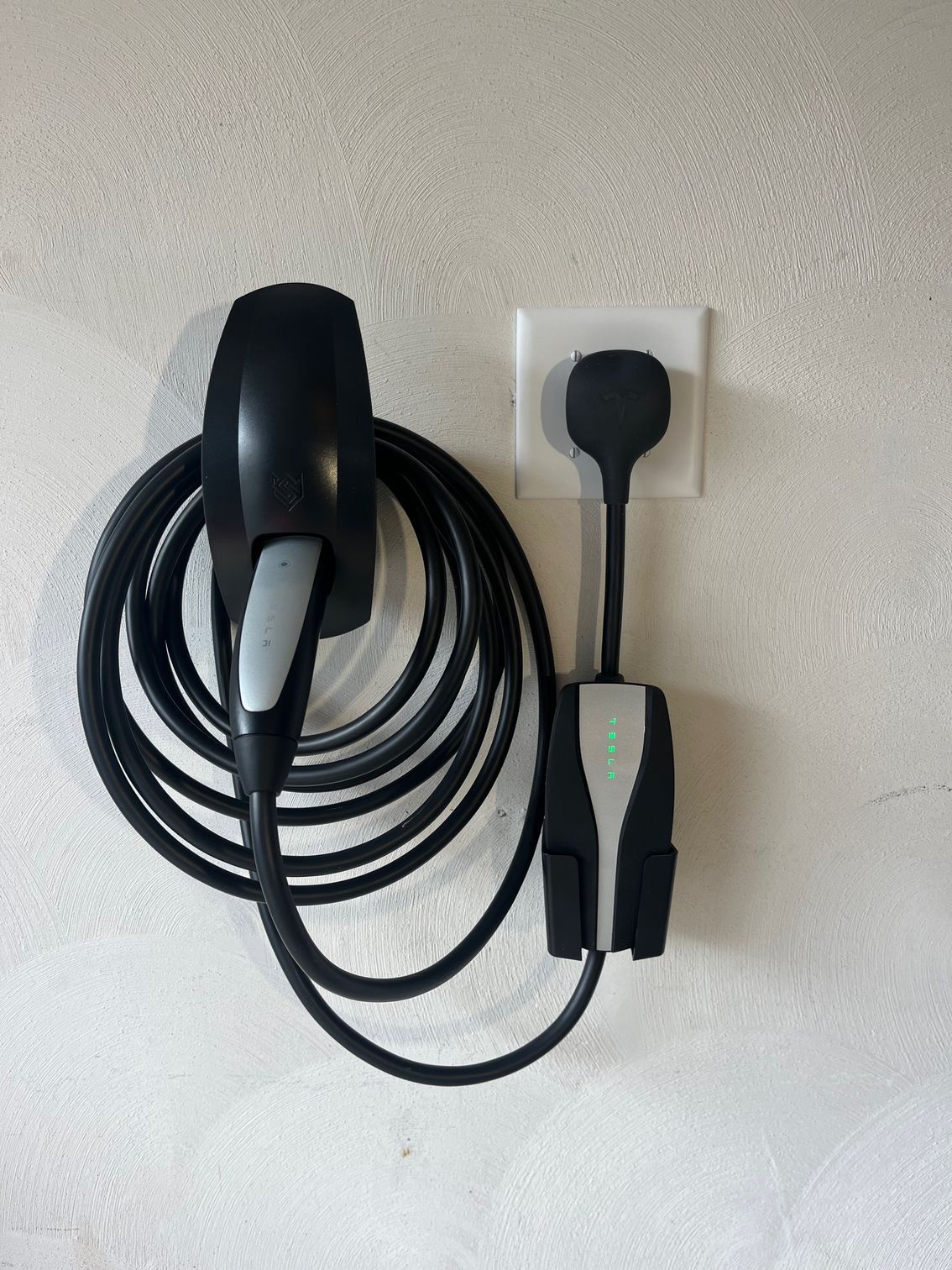 Black EV charger and charging cable coiled on a white wall, plugged into a power outlet.