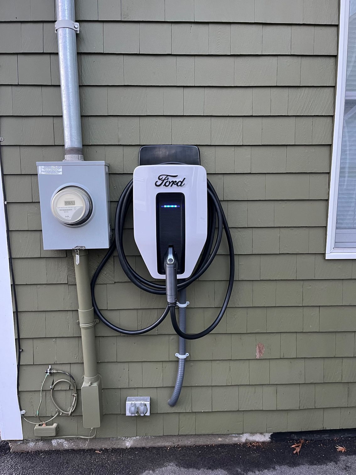 Ford electric vehicle charger mounted on green siding next to electrical meter and window.