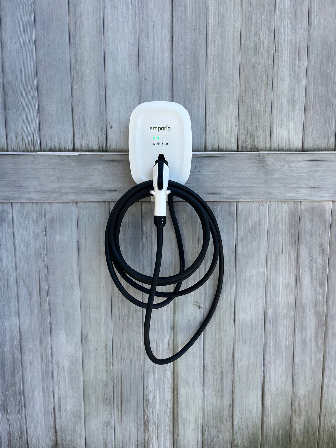 White EV charger mounted on a weathered gray wooden fence. Black charging cable coiled below.
