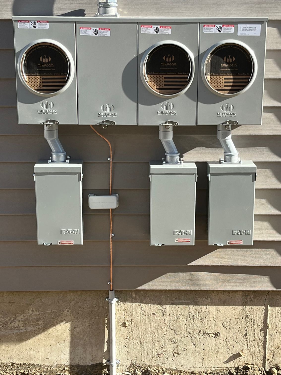 Gray electrical meter box with three units mounted on a brown building exterior.