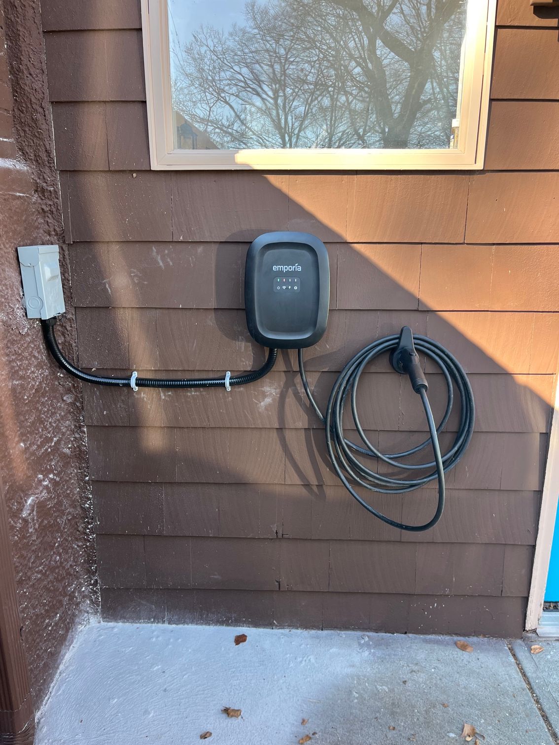 Black EV charger mounted on a brown house exterior next to a window; charging cable coiled below.