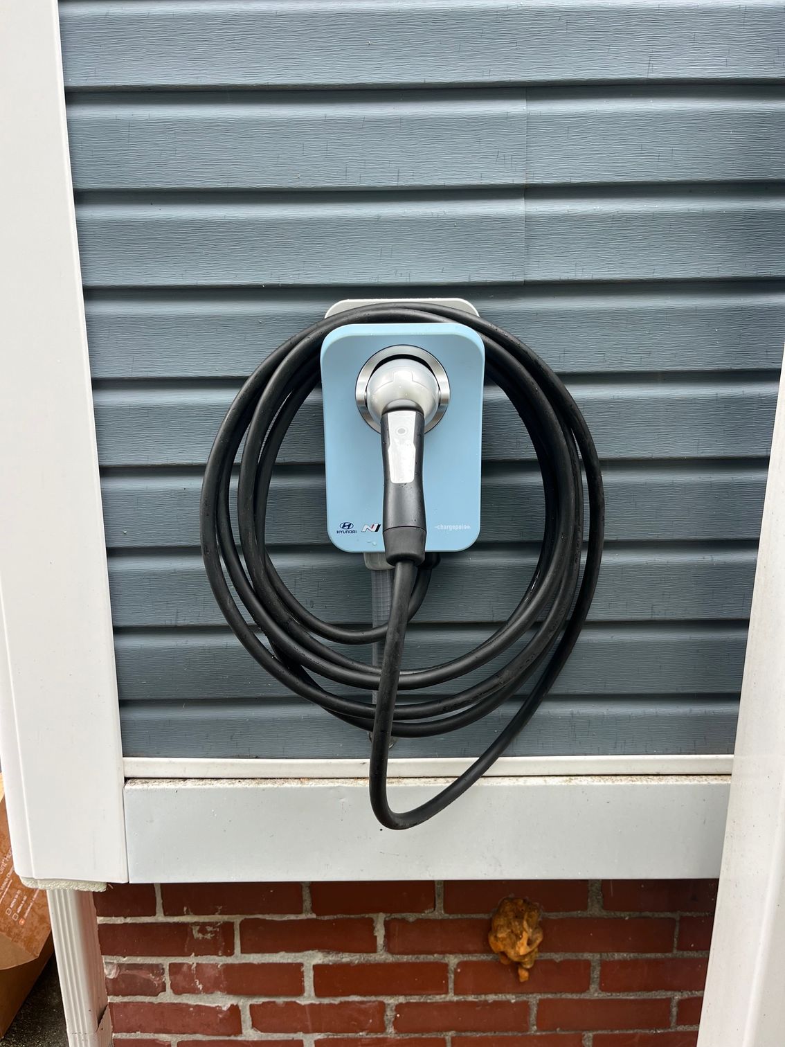 Blue electric vehicle charger mounted on blue siding with black cable coiled.
