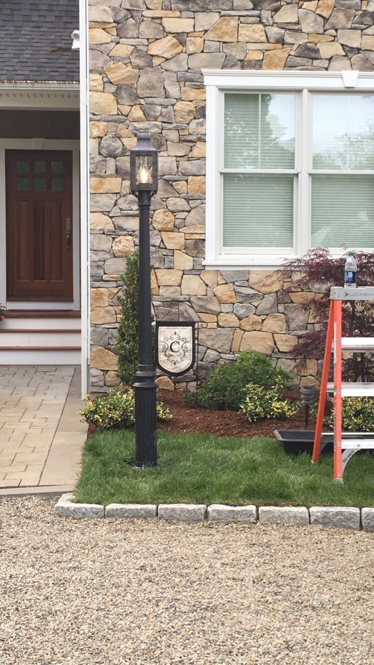 Black lamp post in front of stone house, lit, with sign, grass, and a ladder next to the window.