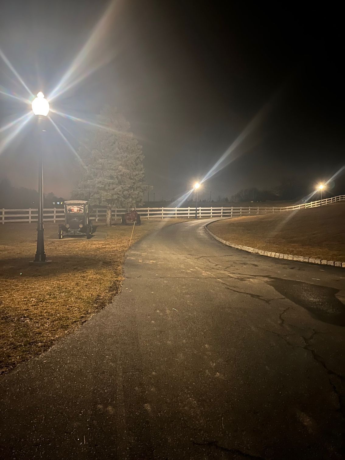 Dark road at night with streetlights illuminating a white fence and classic cars; fog in the background.