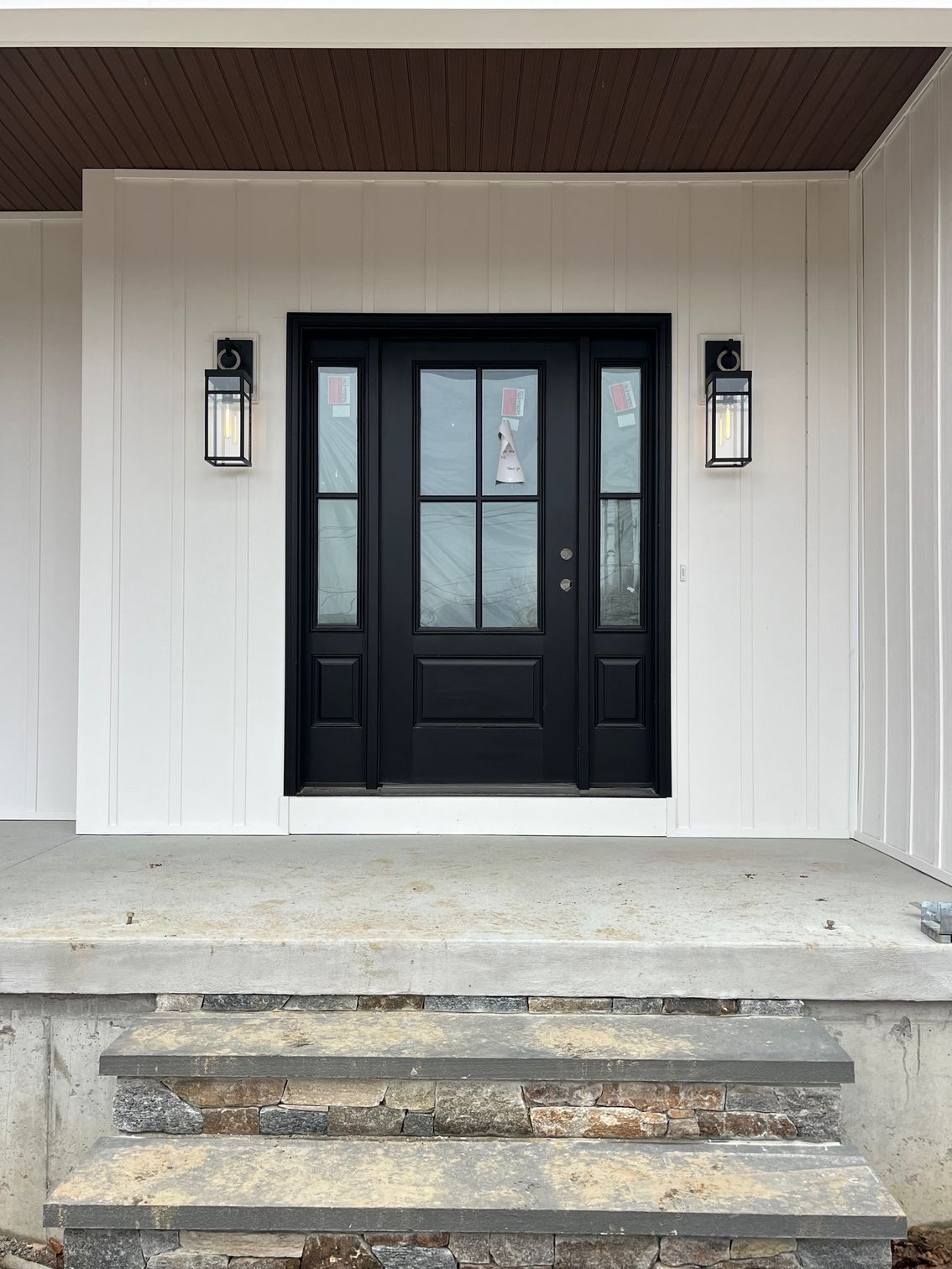 Black front door with sidelights, flanked by sconces, set in white paneling; stone steps lead up.