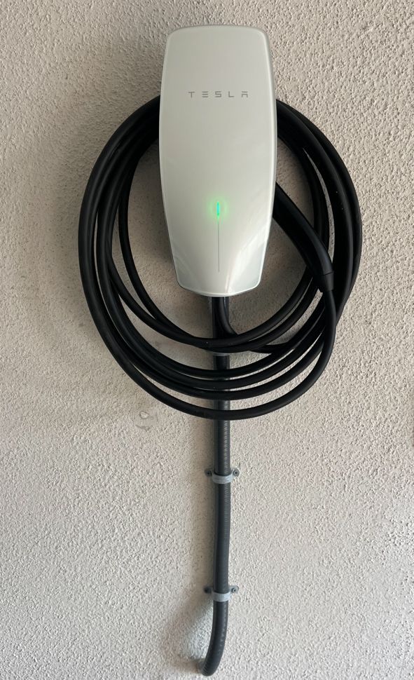 Tesla wall connector with a green light, mounted on a beige wall, cable coiled.