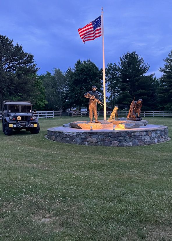 Bronze war memorial with flag, military vehicle, and figures of soldiers and dogs.