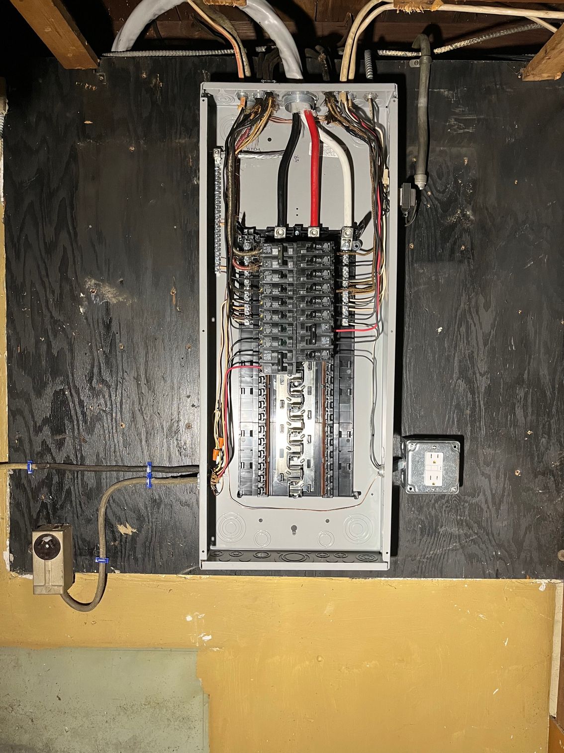 Electrical panel mounted on a dark wooden wall, wires and circuit breakers visible.