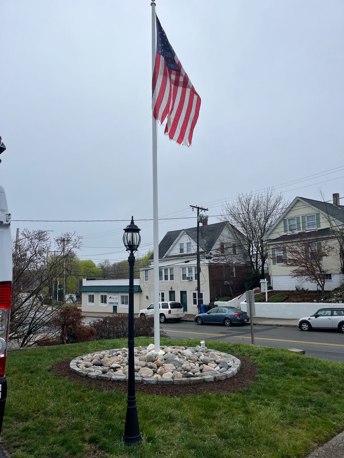 American flag on a flagpole, next to a street with houses and a black lamp post. Cloudy sky.