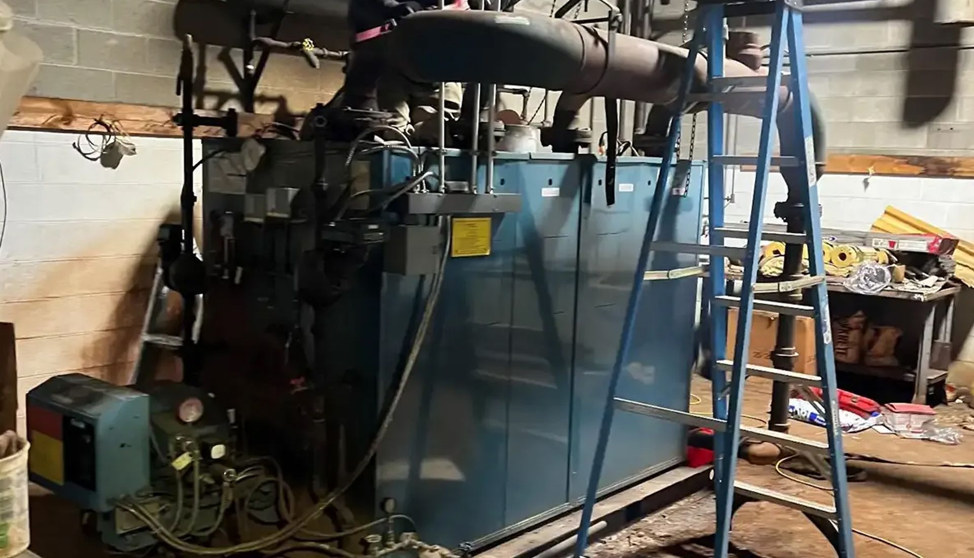 Large blue industrial machine in a cluttered workshop, with a blue ladder leaning against it.