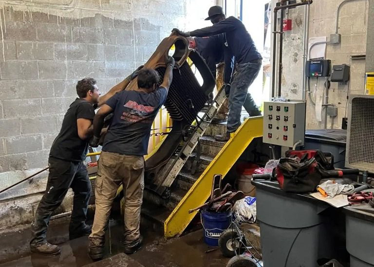Three people in work clothes manually guide a large, dark piece of machinery up a yellow set of industrial stairs.