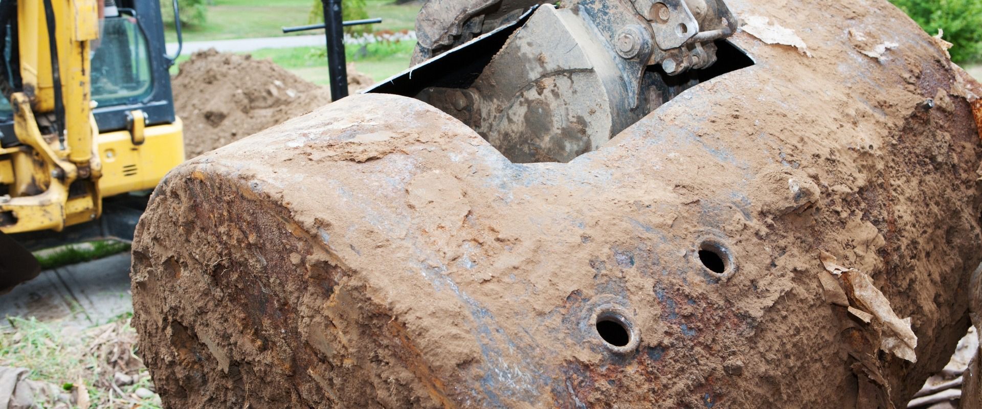 A rusted, excavated underground storage tank sits next to a yellow excavator in a dirt-filled construction area.