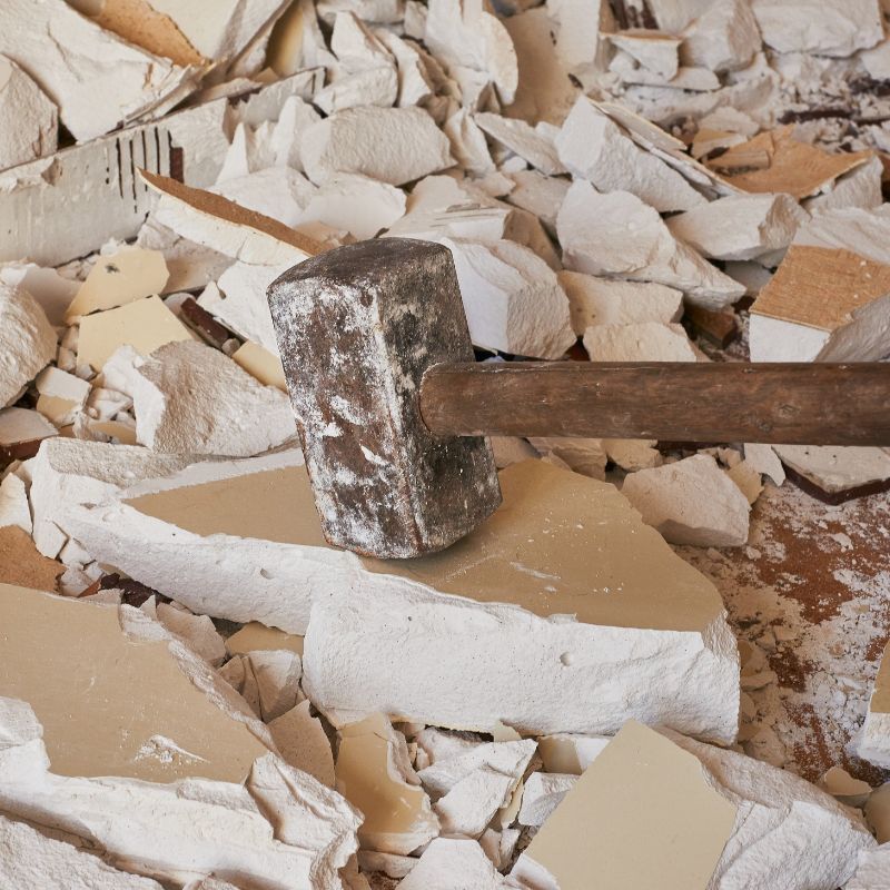 A heavy sledgehammer rests on broken pieces of light-colored drywall amidst construction debris.