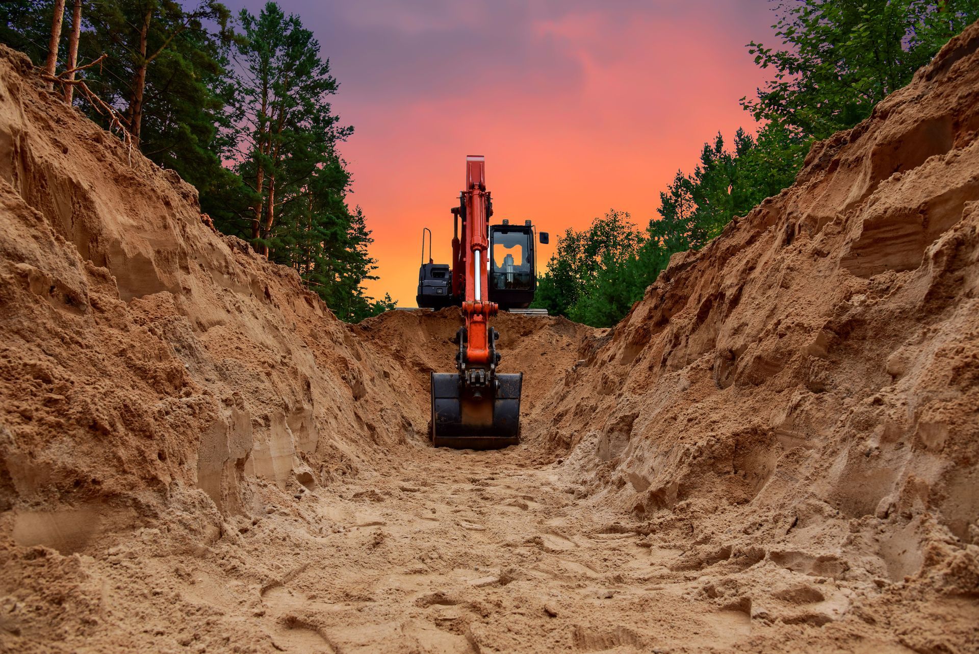 An orange excavator stands inside a deep sandy trench against a vibrant sunset sky.
