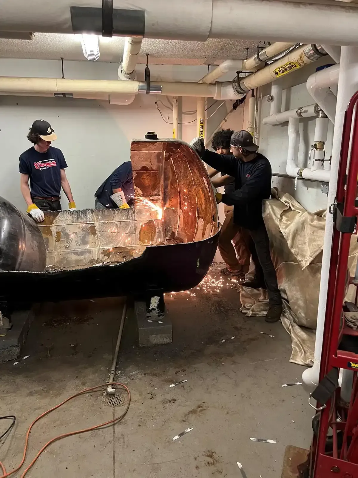 People working on a large rusted pipe in an industrial room, using tools and scaffolding.