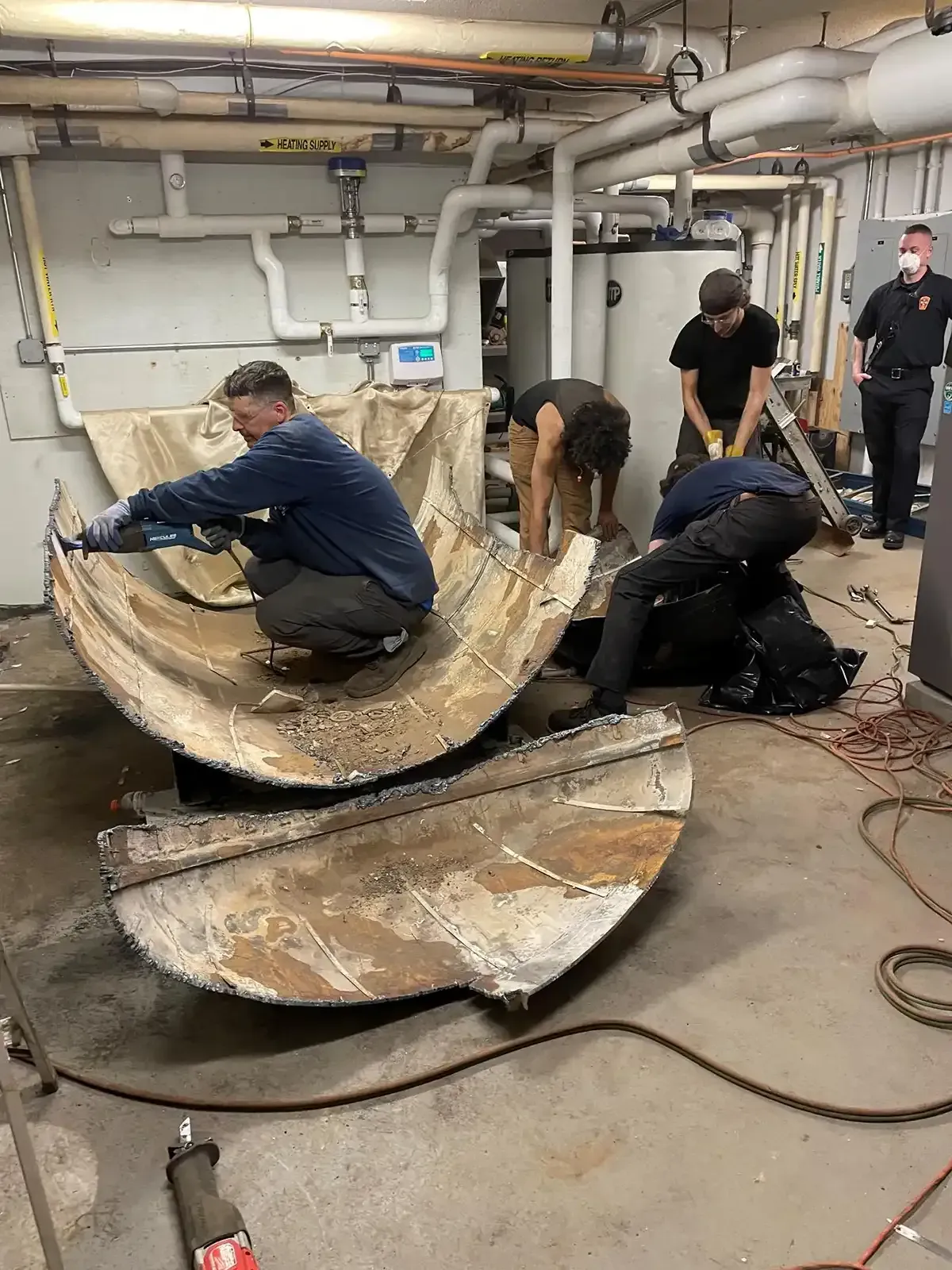 Workers repairing a large broken metal tank in a basement utility room.
