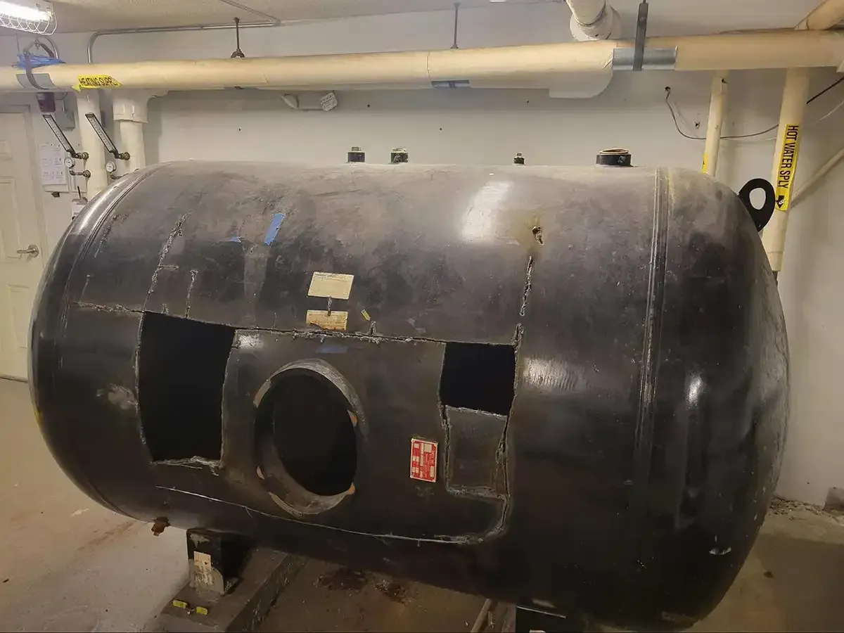 Large black cylindrical tank or vessel in an industrial basement, with openings cut into its side.