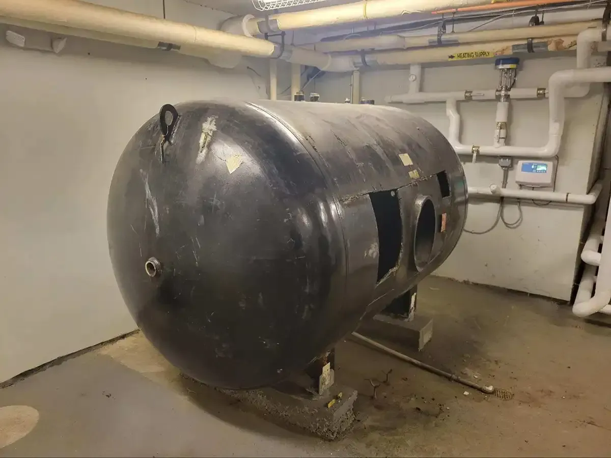 Large black cylindrical tank in a mechanical room with pipes and gauges on the walls.