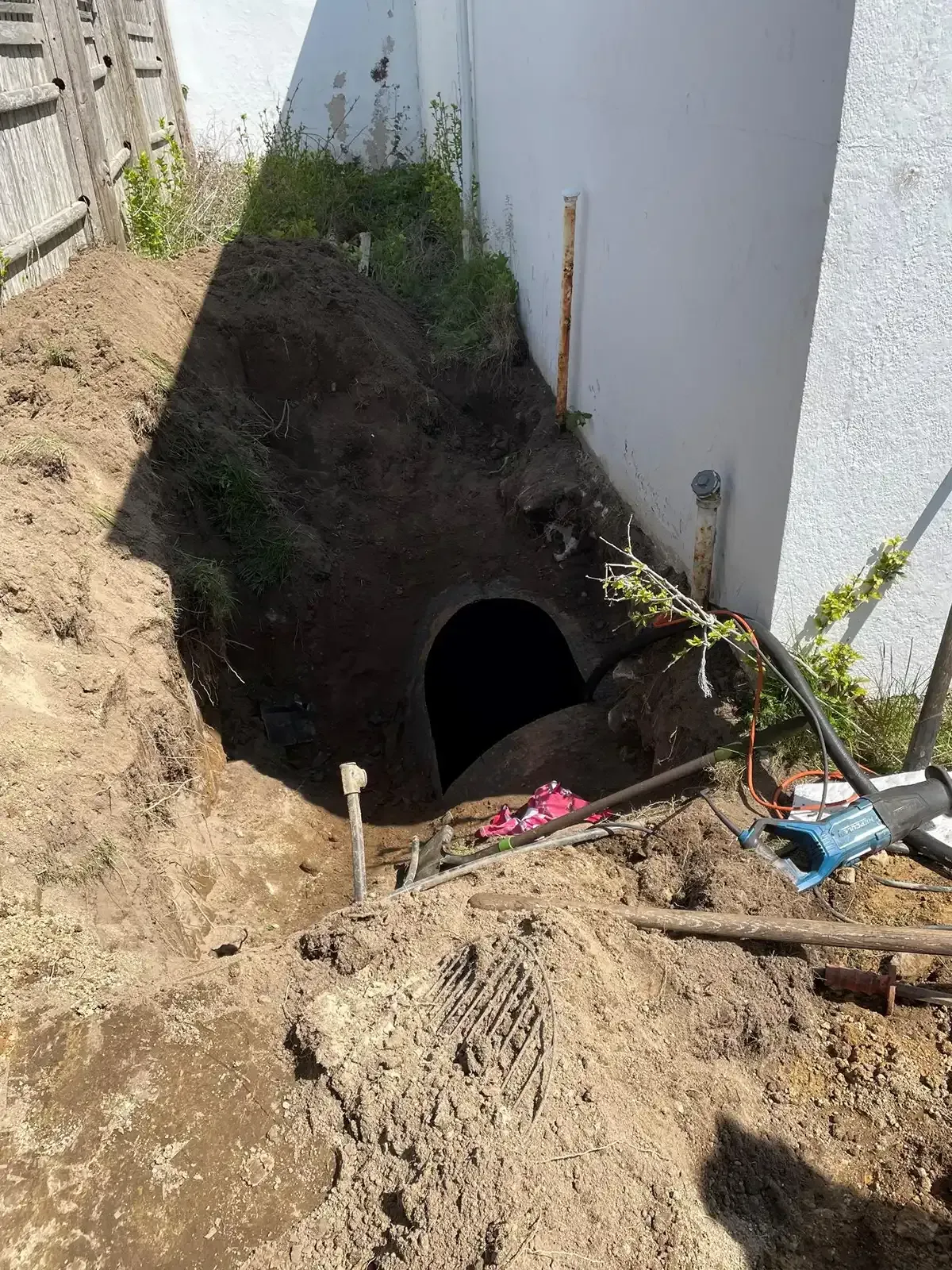 Collapsed trench beside a white wall with a dark tunnel opening and construction tools on the dirt edge.