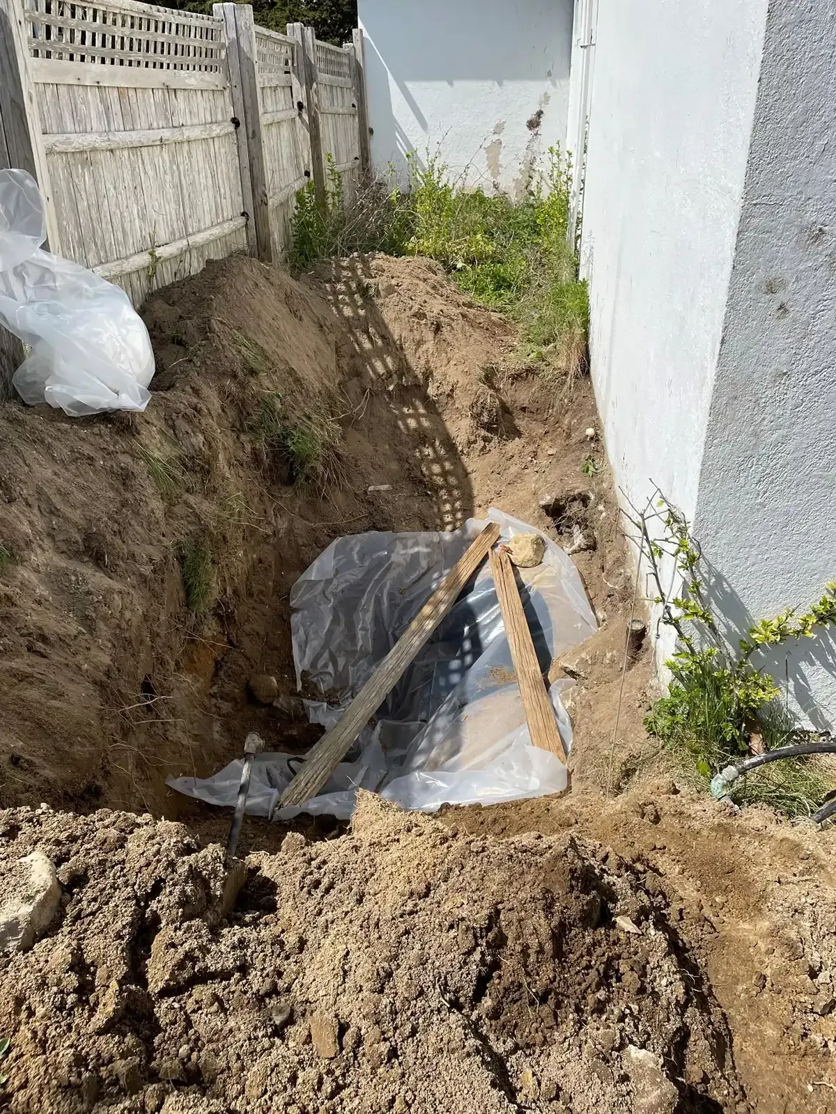 Excavation trench beside a house with dirt piles, black plastic sheeting, and wooden supports.