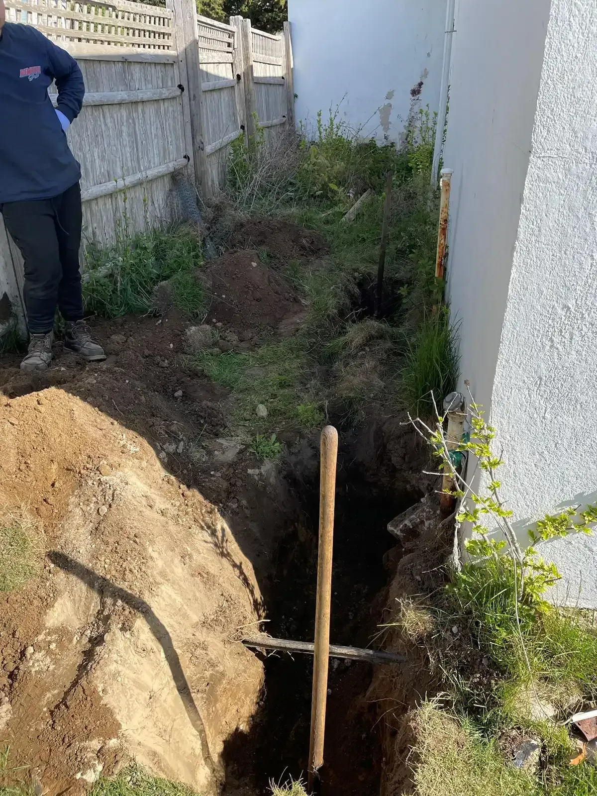 Narrow trench dug beside a white wall, with a shovel and soil piled along the side.