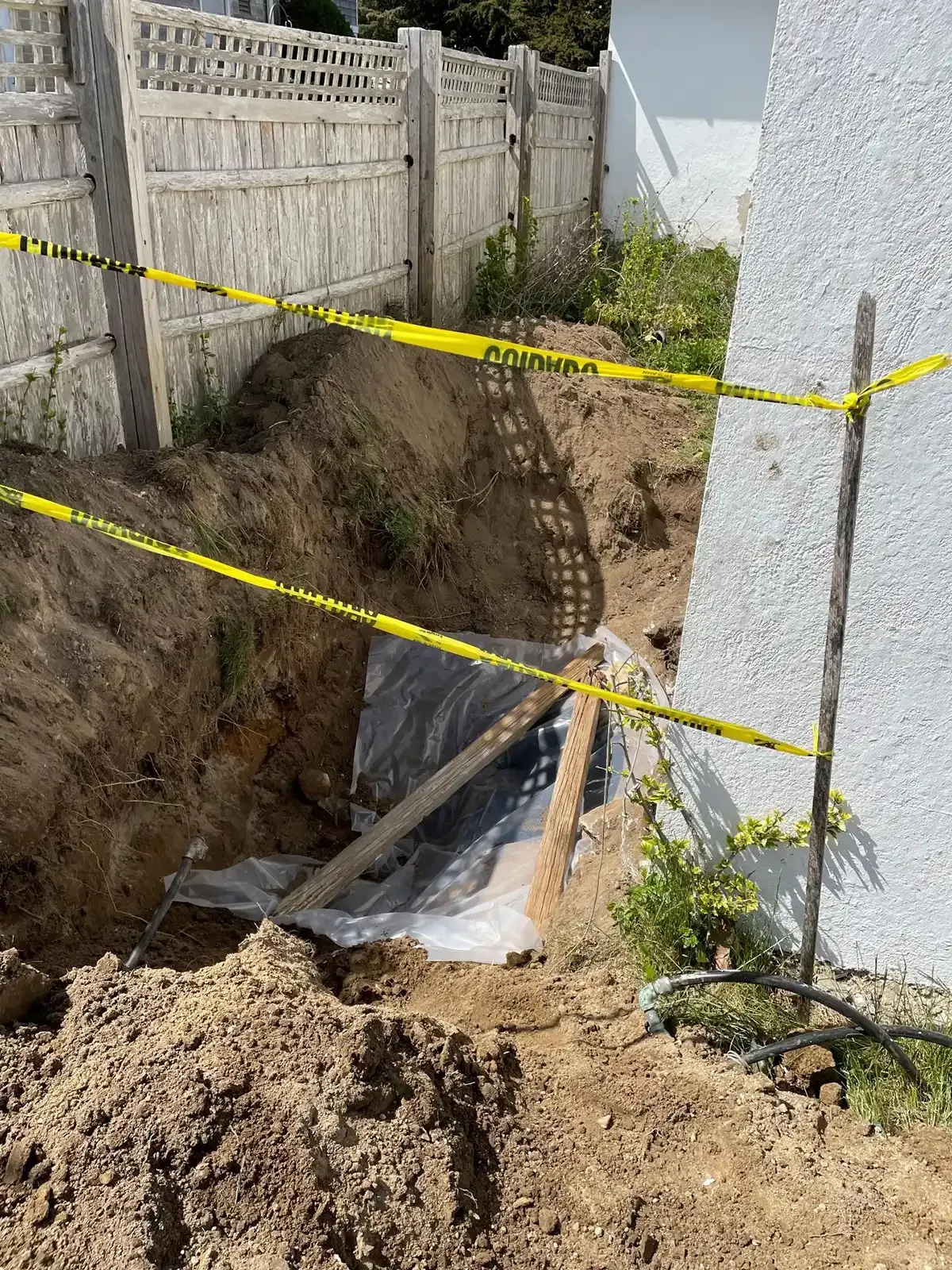 Deep excavation beside a house, cordoned with yellow caution tape and dirt piles around a trench.