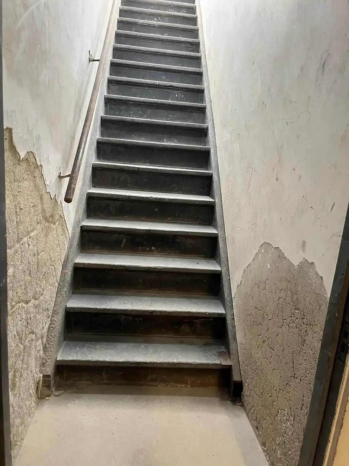 Narrow concrete stairwell with worn white walls and steep steps leading upward.