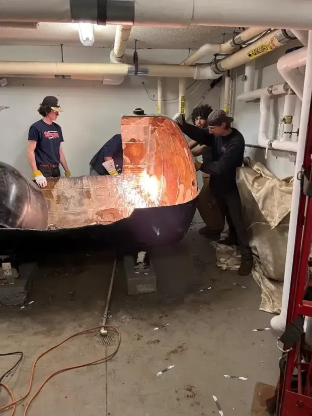 Three people in a mechanical room work on a large, partially dismantled purple metal object with exposed copper interior.