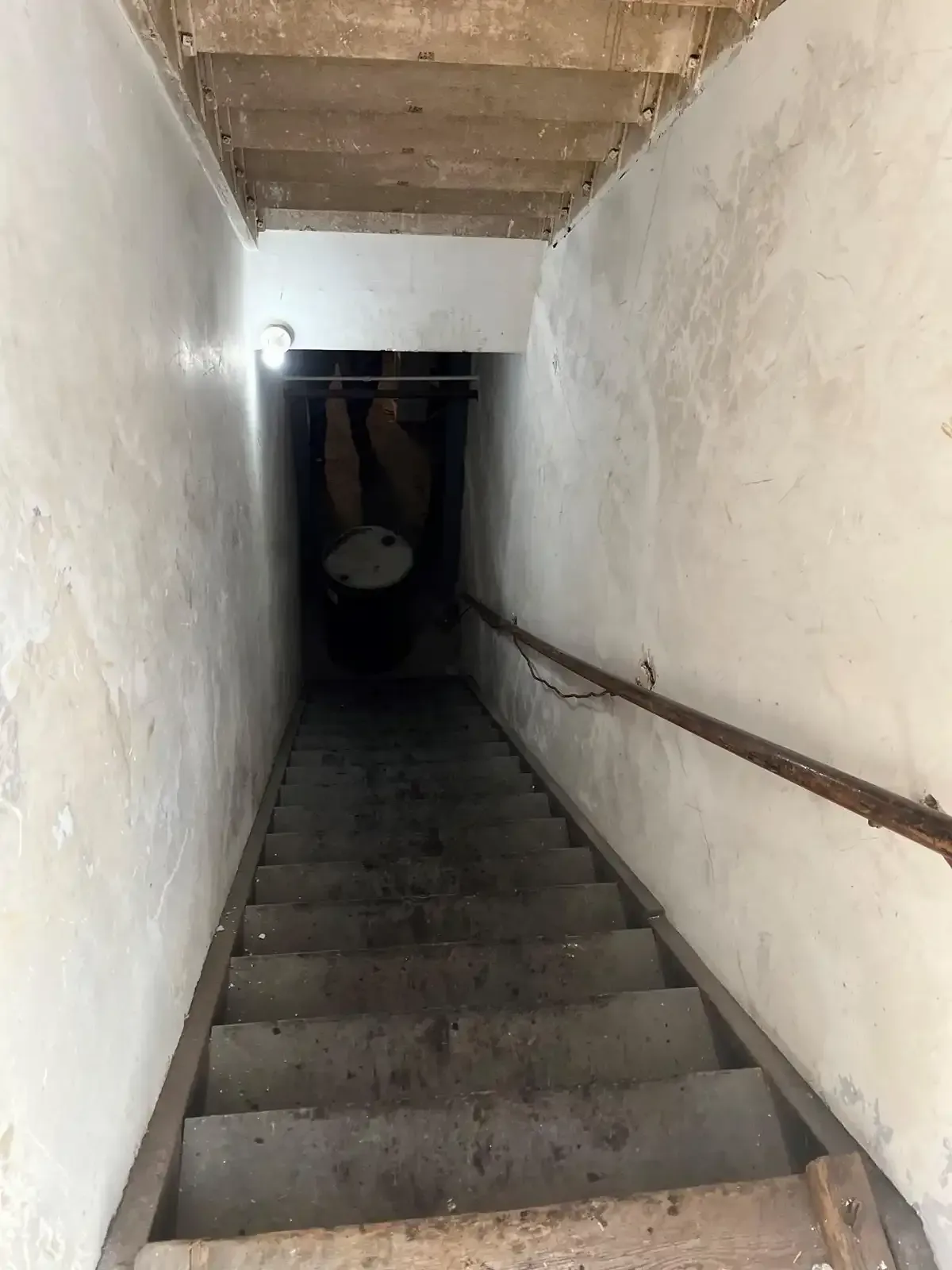 Narrow concrete stairwell descending into a dark basement with white walls and a handrail.