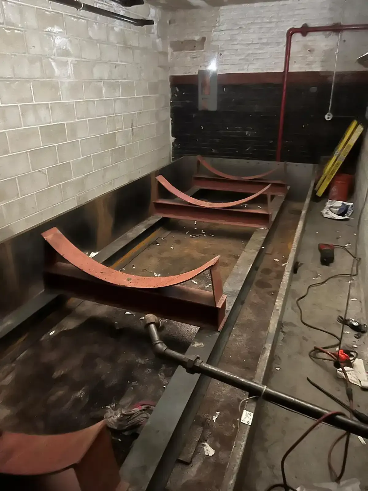 Industrial kitchen floor with metal-framed equipment under construction, pipes and tools along the wall.