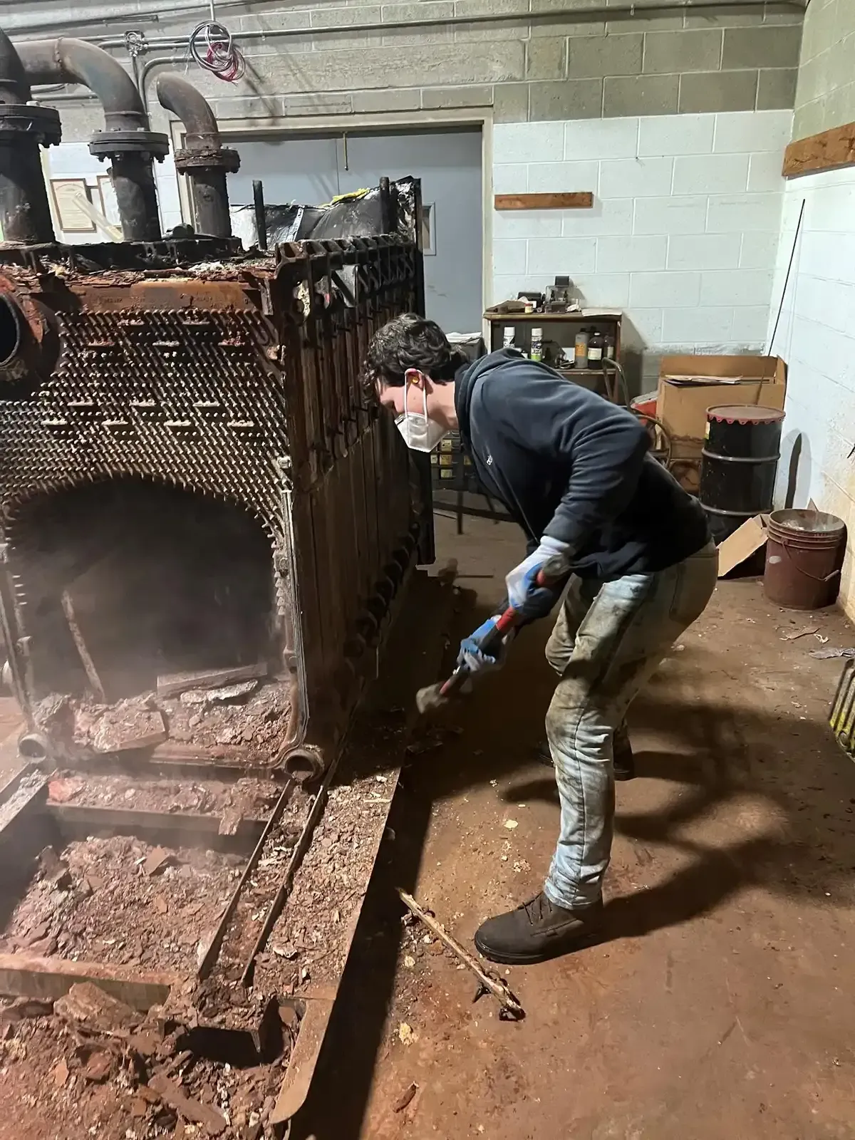 Person cleaning rust from a large industrial boiler in a basement utility room.