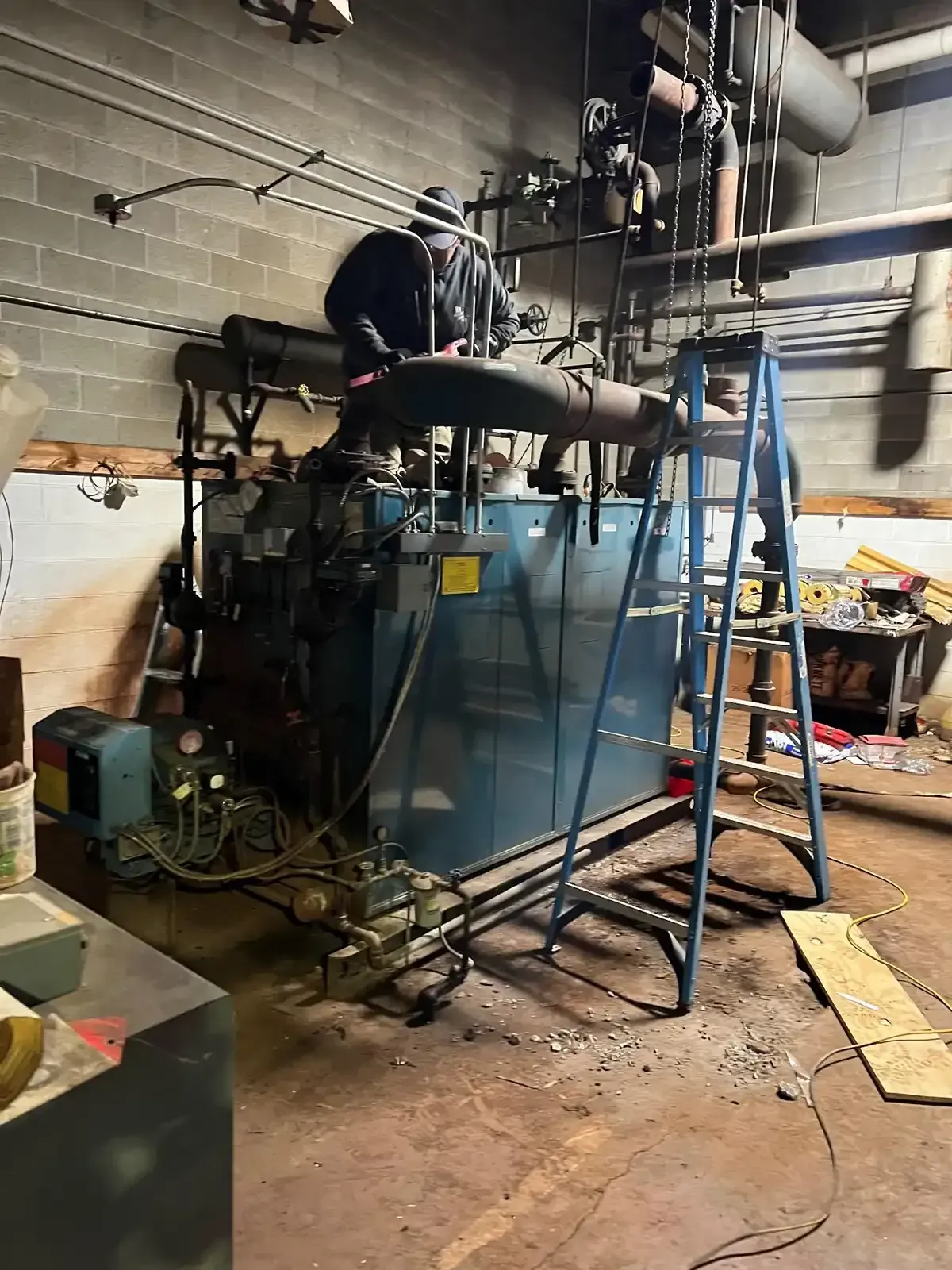 Large blue industrial machine in a cluttered workshop, with ladders, pipes, and tools around it.