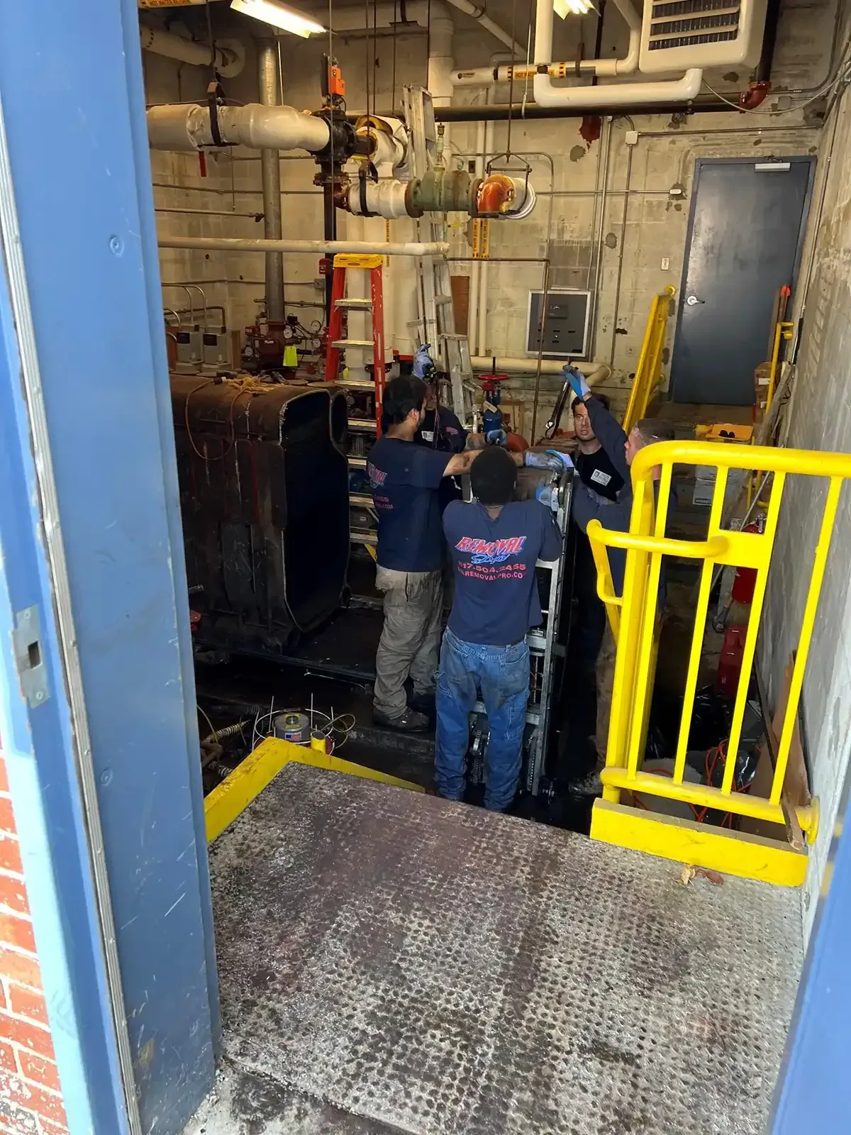 Workers in a utility pit with pipes and a yellow safety railing in an industrial room.