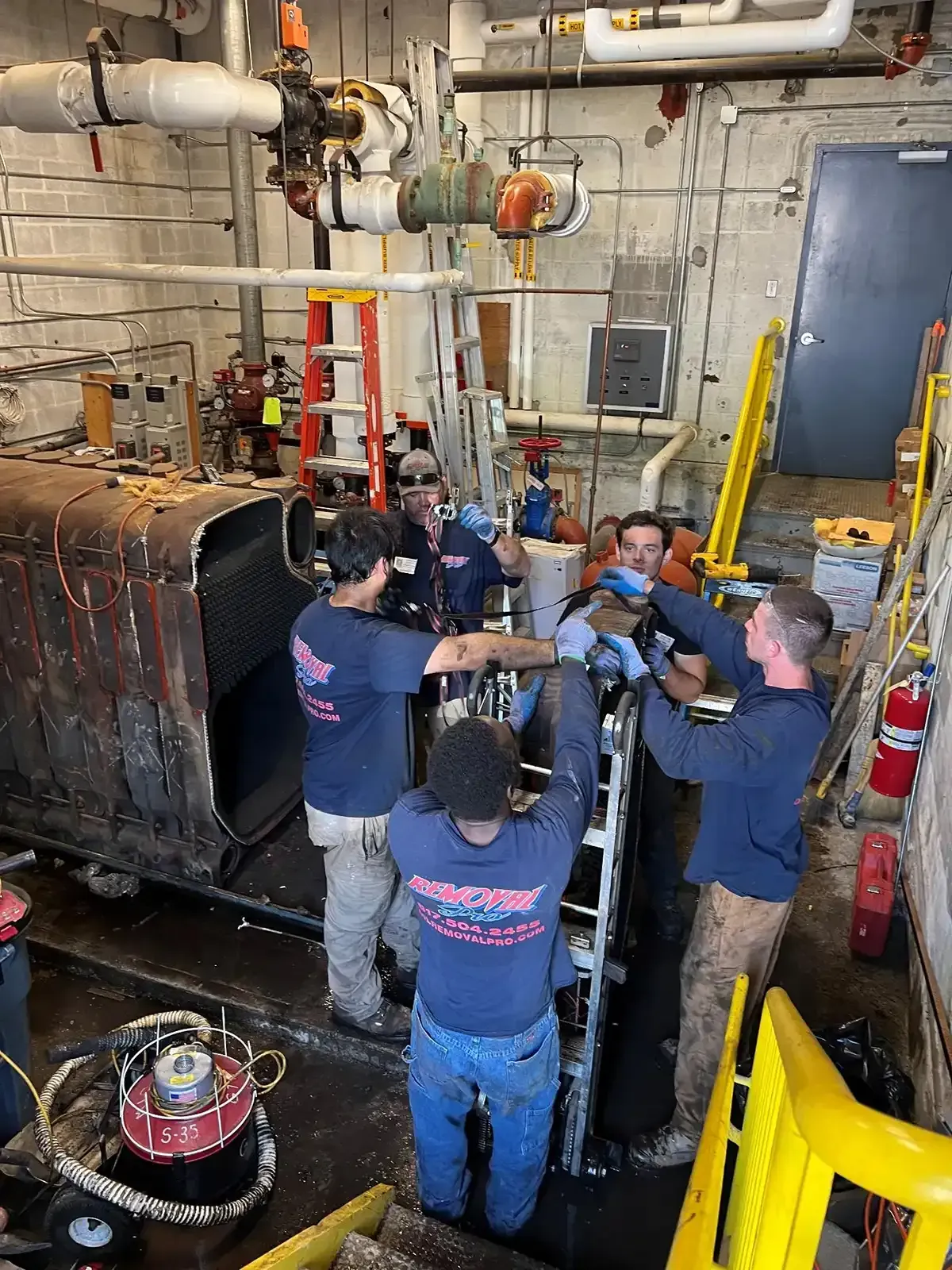 Workers inspecting machinery in an industrial room with pipes, valves, and a large tank.