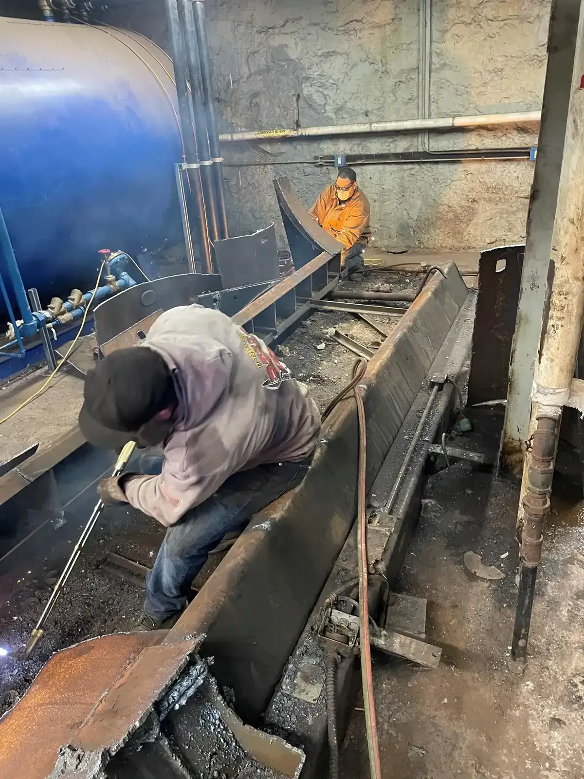 Two workers in a narrow industrial trench, one crouching by a pipe and one in an orange jacket nearby.