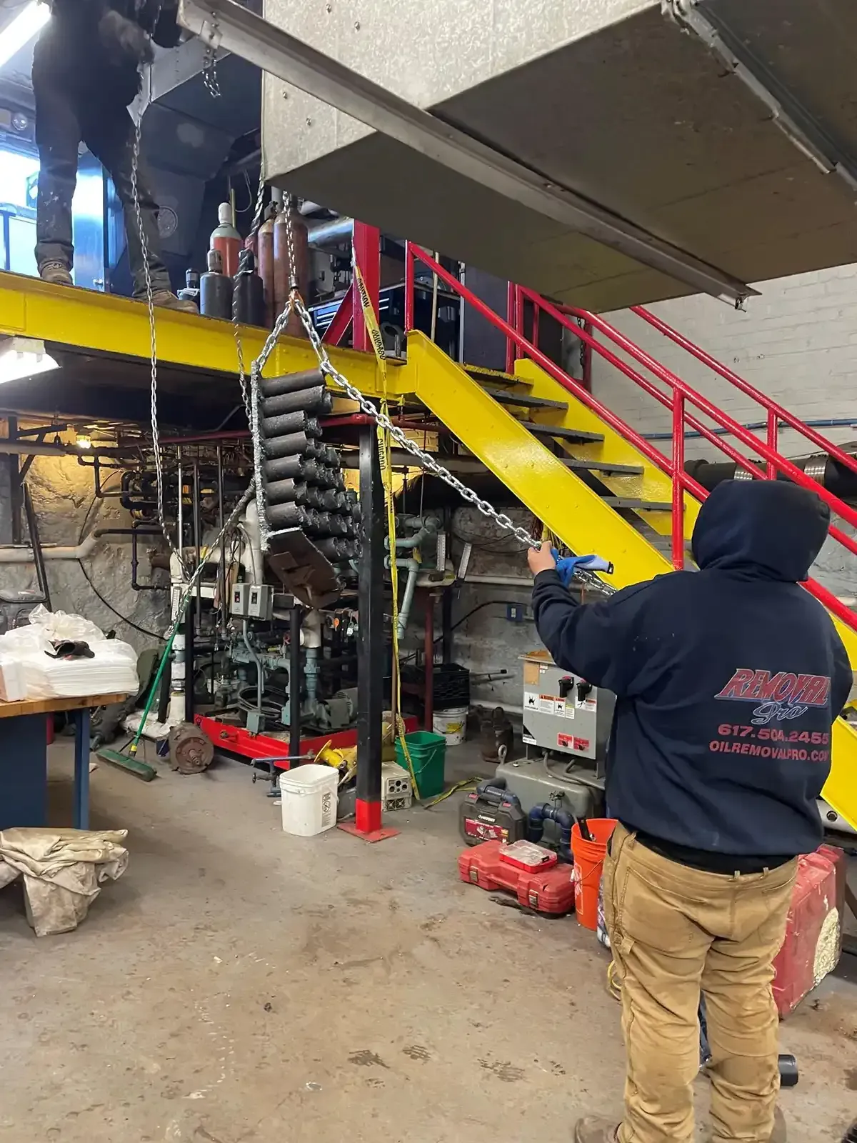 Worker in navy hoodie points at a yellow staircase and red railings in an industrial workspace.