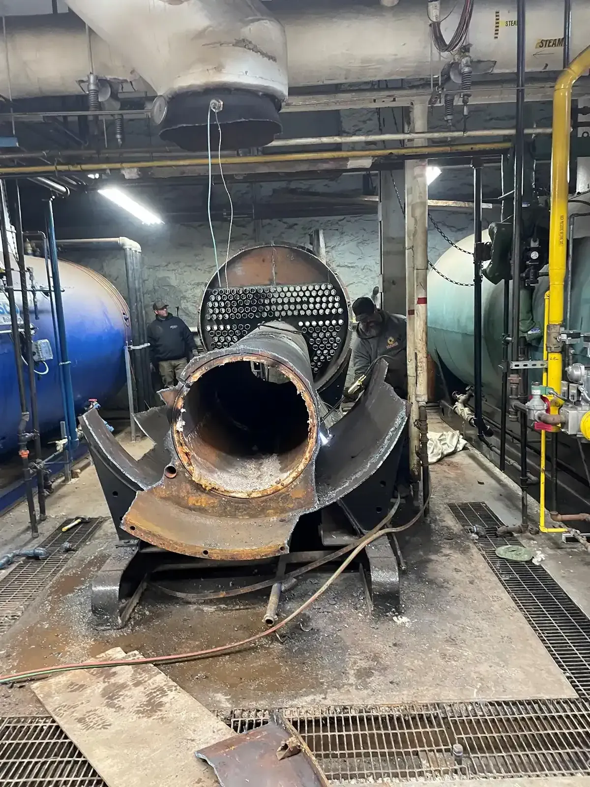Damaged industrial tank with large open pipe in a wet factory room, debris and equipment around it.